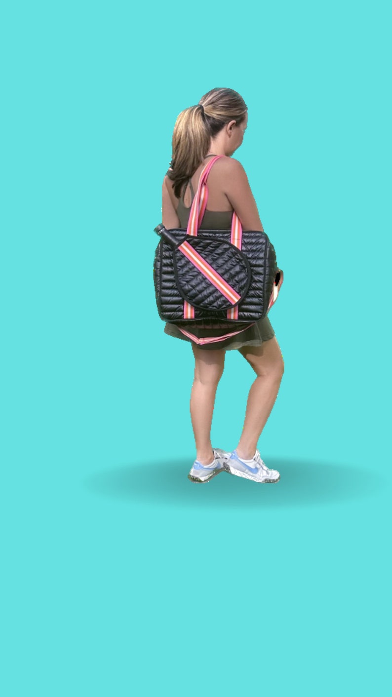 Puede incluir: Una bolsa de tenis acolchada negra con rayas rosas y blancas. La bolsa tiene un soporte para raquetas y un compartimento con cremallera. La bolsa la lleva una persona que lleva un top verde y una camiseta blanca.
