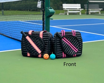 May include: Two black quilted bags with pink and white straps. Each bag has a paddle racket inside and a pocket on the front. Two orange and blue pickleball balls are in front of the bags. The text "Front" is written below the bags.