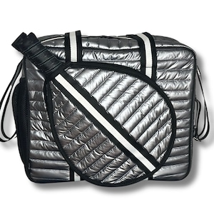 Puede incluir: Una bolsa de viaje acolchada plateada con una correa a rayas blancas y negras y un porta raquetas acolchado blanco y negro.