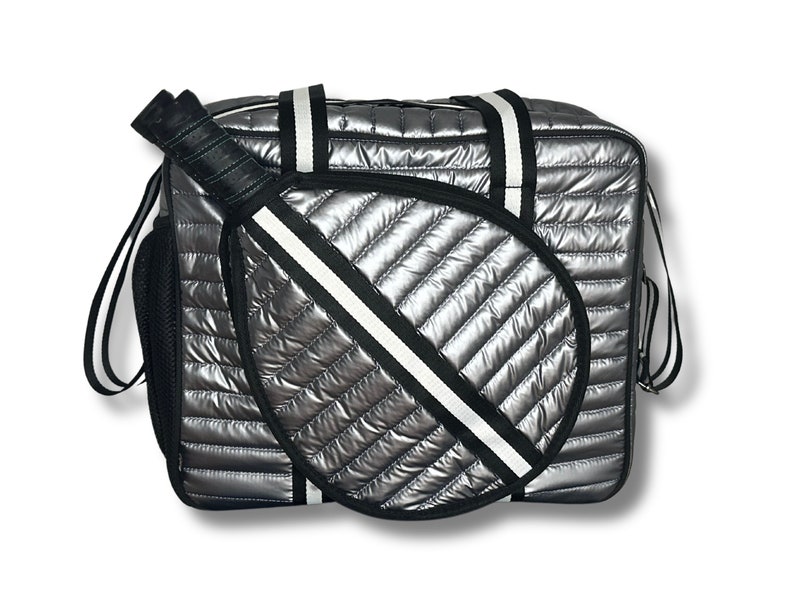 Puede incluir: Una bolsa de viaje acolchada plateada con una correa a rayas blancas y negras y un porta raquetas acolchado blanco y negro.