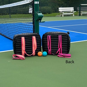 Puede incluir: Dos bolsas acolchadas negras con correas rosas y naranjas, y dos pelotas de pickleball en una cancha de pickleball azul y verde. El texto "Back" es visible en la cancha.