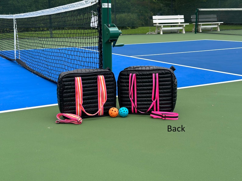 Puede incluir: Dos bolsas acolchadas negras con correas rosas y naranjas, y dos pelotas de pickleball en una cancha de pickleball azul y verde. El texto "Back" es visible en la cancha.