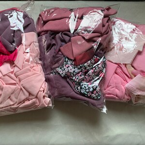 May include: Three clear plastic bags filled with assorted fabric scraps in various shades of pink, magenta, and mauve. The collection includes solid-coloured textiles and one piece featuring a black fabric with a small white and pink floral pattern.