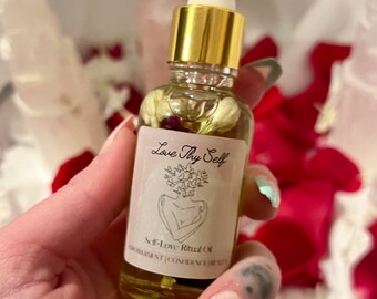 May include: A small glass bottle of Self-Love Ritual Oil with a gold cap and white dropper. The label reads "Love Thy Self" with an illustration. The oil contains flowers and is held against a background of red and white petals.