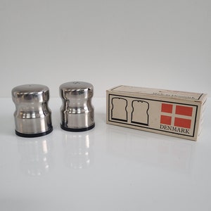 May include: Two stainless steel salt and pepper shakers with a black base, next to a box with the text "Made in Denmark" and the Danish flag. The box features illustrations of two slices of bread.