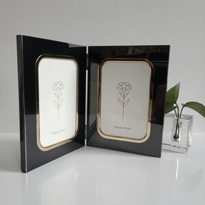 May include: A black double picture frame with gold trim, designed to hold two 4" x 6" photos. Each frame features a white background with a floral line drawing. The frame is hinged and stands upright.
