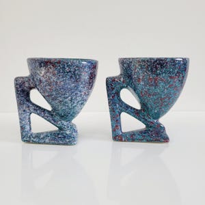 May include: Two speckled ceramic cups with geometric handles. The cups have a blue and green speckled pattern with red accents. The cups are displayed on a white surface.
