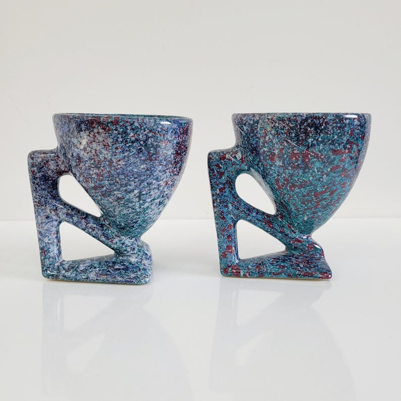 May include: Two speckled ceramic cups with geometric handles. The cups have a blue and green speckled pattern with red accents. The cups are displayed on a white surface.