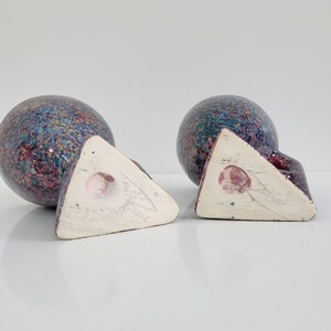 May include: Two ceramic bookends with a speckled blue, purple, and red pattern. Each bookend has a spherical shape and a triangular base. The bases are white with a pink floral design.
