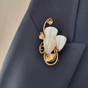 May include: A gold-toned brooch in the shape of a flower with three petal-shaped stones. The brooch is attached to a navy blue blazer. The petals are white and beige, and the brooch has a detailed, ornate design.