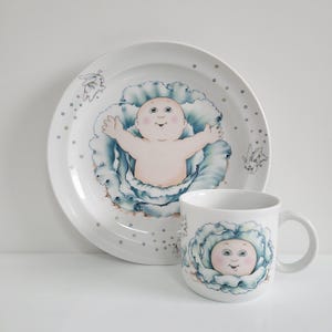 May include: A white ceramic plate and mug set. The plate features a baby design surrounded by a blue cabbage-like shape. The mug has a similar design. Both pieces have a whimsical, hand-painted style.