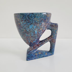 May include: A speckled ceramic mug with a unique handle design. The mug features a blue, red, and gold speckled pattern. The handle has a geometric cutout design. The mug is a decorative piece.