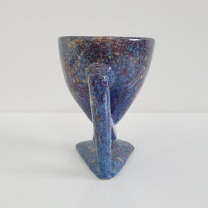 May include: A speckled ceramic cup with a unique handle and base. The cup features a blue background with red, gold, and brown speckles. The handle is a curved, elongated shape, and the base is triangular.