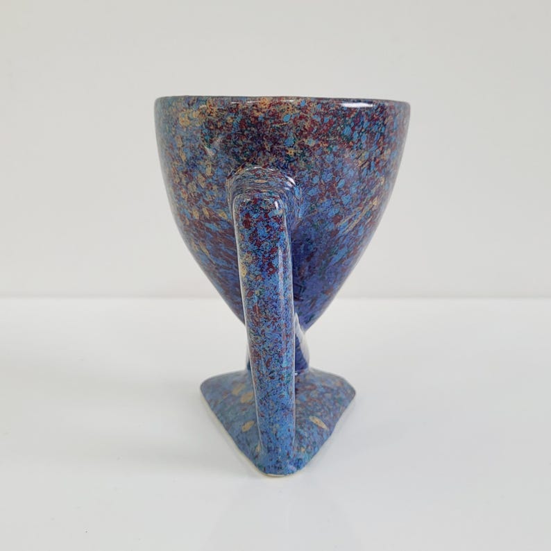 May include: A speckled ceramic cup with a unique handle and base. The cup features a blue background with red, gold, and brown speckles. The handle is a curved, elongated shape, and the base is triangular.