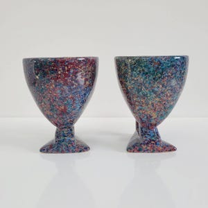 May include: Two speckled ceramic goblets. The goblets have a wide bowl shape and a pedestal base. The color scheme is a mix of blue, red, and yellow speckles. The goblets are likely for decorative use or serving beverages.