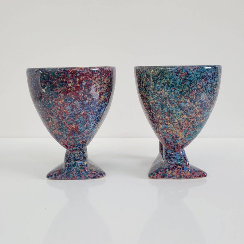 May include: Two speckled ceramic goblets. The goblets have a wide bowl shape and a pedestal base. The color scheme is a mix of blue, red, and yellow speckles. The goblets are likely for decorative use or serving beverages.