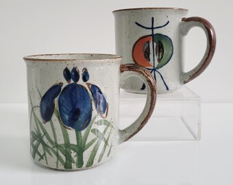 May include: Two ceramic mugs with speckled, off-white glaze. One mug features a blue iris flower design with green leaves. The other mug has an abstract design in orange, green, and blue. Both mugs have brown handles and rims.