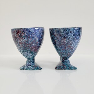 May include: Two speckled blue and maroon ceramic goblets with a flared top and pedestal base. The goblets have a speckled pattern with shades of blue, teal, and maroon. The base is a darker teal color.