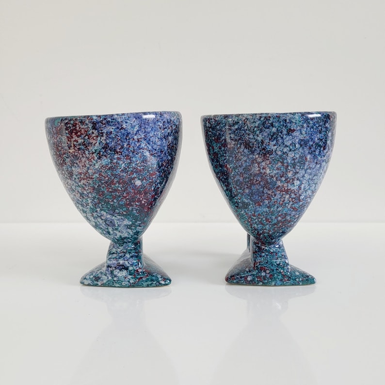May include: Two speckled blue and maroon ceramic goblets with a flared top and pedestal base. The goblets have a speckled pattern with shades of blue, teal, and maroon. The base is a darker teal color.