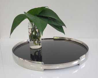 May include: A round serving tray with a reflective black surface and a polished silver-tone rim. A small glass jar with green leaves and stems sits on the tray. The tray is a decorative item for serving or displaying items.