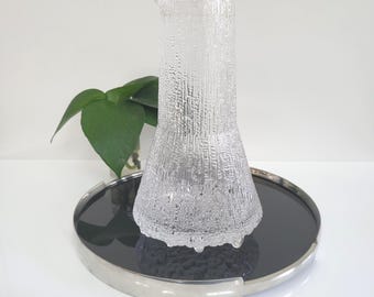 May include: A clear glass pitcher with a textured, bark-like pattern. The pitcher sits on a black, circular tray with a silver rim. A green plant is visible in the background.