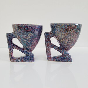 May include: Two speckled ceramic cups with geometric handles. The cups have a unique design with a speckled pattern in shades of blue, purple, and red. The handles are angular and add to the modern aesthetic. The cups are displayed on a white surface.