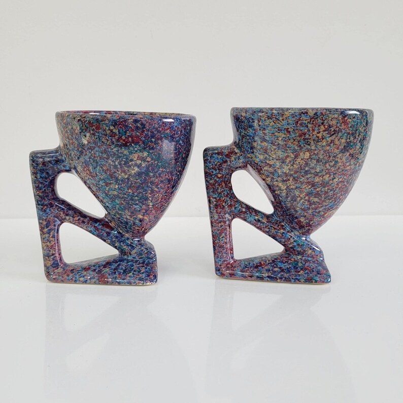 May include: Two speckled ceramic cups with geometric handles. The cups have a unique design with a speckled pattern in shades of blue, purple, and red. The handles are angular and add to the modern aesthetic. The cups are displayed on a white surface.