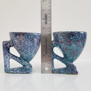 May include: Two speckled ceramic mugs with a unique handle design. The mugs are primarily blue with red and green accents. A stainless steel ruler is placed between the mugs, showing their height. The mugs have a modern, artistic aesthetic.
