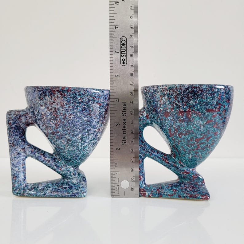 May include: Two speckled ceramic mugs with a unique handle design. The mugs are primarily blue with red and green accents. A stainless steel ruler is placed between the mugs, showing their height. The mugs have a modern, artistic aesthetic.