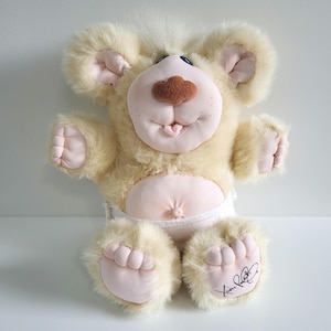 May include: A plush bear toy with a light tan fur body, large ears, and pink accents on the paws, ears, and face. The bear has a brown nose, blue eyes, and a smiling expression. It wears a white nappy and has a signature on its foot.