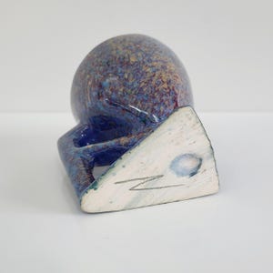 May include: A handmade ceramic mug with a unique design. The mug features a speckled blue and purple exterior and a triangular base. The handle is integrated into the design, and the base has a white surface with a signature.