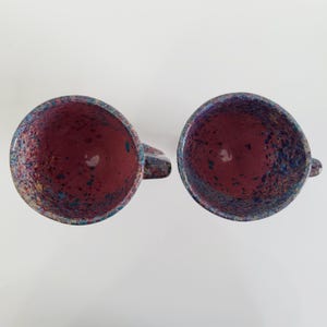 May include: Two ceramic mugs with a speckled design. The interior of each mug is a deep burgundy color, while the exterior rim and handle feature a mix of blue, purple, and yellow speckles. The mugs are set against a white background.