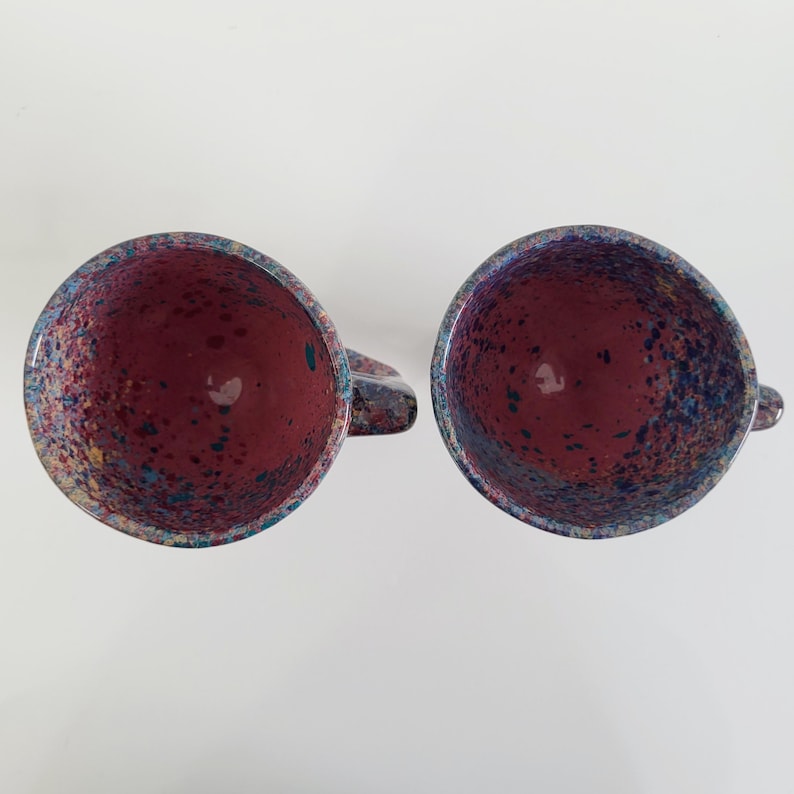 May include: Two ceramic mugs with a speckled design. The interior of each mug is a deep burgundy color, while the exterior rim and handle feature a mix of blue, purple, and yellow speckles. The mugs are set against a white background.