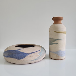 Peut inclure: Un vase en céramique à col étroit et un bol en céramique bas et large. Les deux pièces présentent une base texturée blanc cassé avec des éclaboussures abstraites de bleu, bleu clair et jaune sourd. Le vase a un bord en terre cuite.