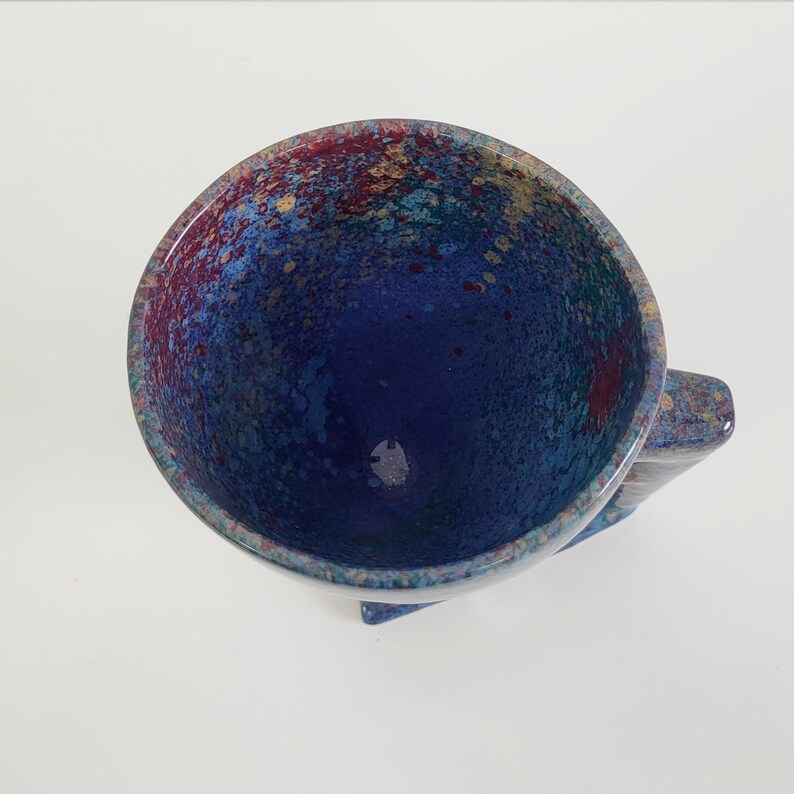 May include: A handmade ceramic mug with a speckled, colorful interior. The interior features a deep blue base with splashes of red, gold, and teal. The exterior handle and base are a muted blue with speckled accents. A unique piece for coffee or tea.