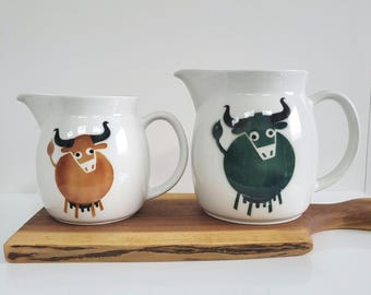 May include: Two white ceramic pitchers with bull illustrations. One pitcher features a brown bull, the other a green bull. Both have black horns and legs. The pitchers sit on a wooden cutting board.