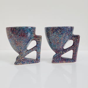 May include: Two speckled ceramic cups with a unique design. The cups have a flared top, a handle with a geometric cutout, and a speckled pattern in shades of blue, red, and yellow. The cups are on a white surface.