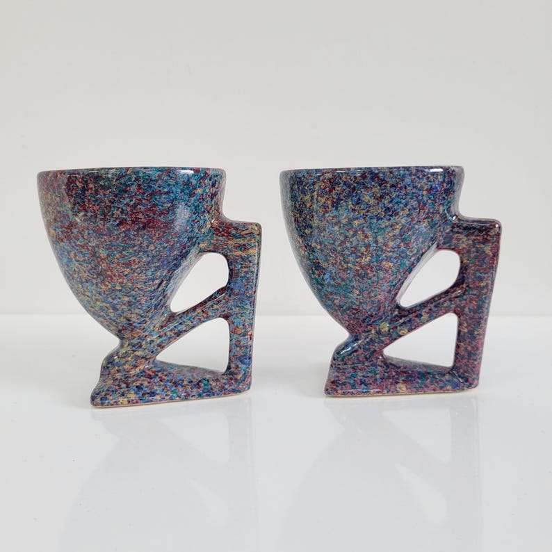 May include: Two speckled ceramic cups with a unique design. The cups have a flared top, a handle with a geometric cutout, and a speckled pattern in shades of blue, red, and yellow. The cups are on a white surface.
