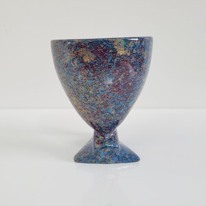 May include: A speckled ceramic goblet with a wide bowl and a triangular base. The goblet features a blend of blue, red, and gold colors. The stem is short and the base is a solid blue.