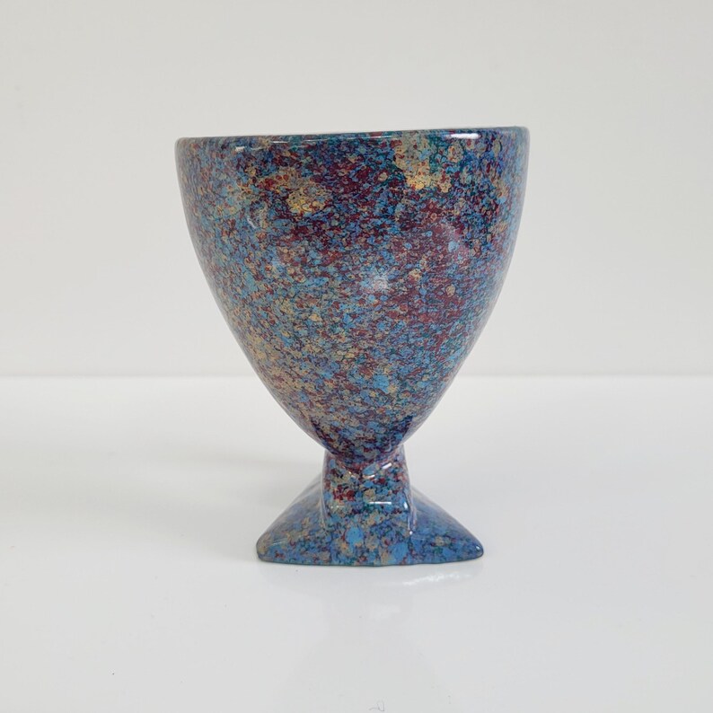 May include: A speckled ceramic goblet with a wide bowl and a triangular base. The goblet features a blend of blue, red, and gold colors. The stem is short and the base is a solid blue.