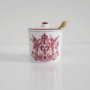 Könnte beinhalten: Weißer Keramiktopf mit Deckel und kleinem Holzlöffel. Der Topf hat ein rotes Wappen-Design mit dem Text "QUE MAILLE QUI MAILLE". Der Rand und die Oberseite des Deckels sind ebenfalls rot.