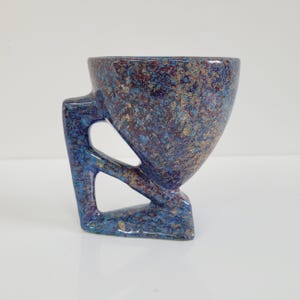 May include: A speckled ceramic cup with a unique handle design. The cup features a blue, brown, and gold speckled pattern. The handle has an open, geometric design, and the cup sits on a small base.