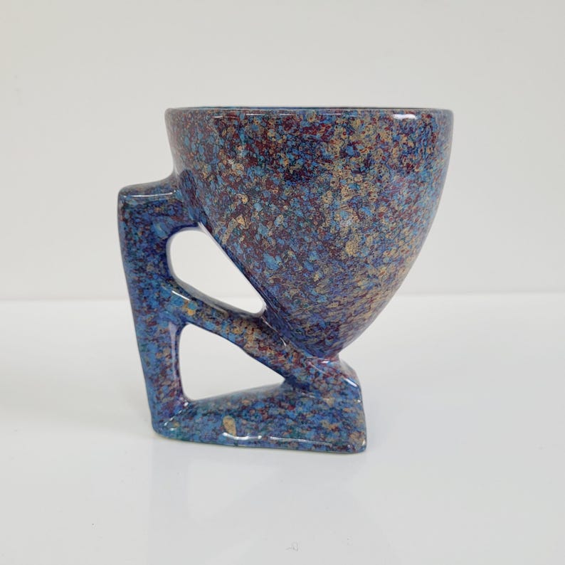 May include: A speckled ceramic cup with a unique handle design. The cup features a blue, brown, and gold speckled pattern. The handle has an open, geometric design, and the cup sits on a small base.