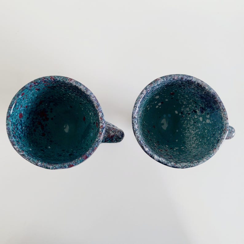 May include: Two ceramic mugs with a speckled teal glaze and small handles. The interior of the mugs is a solid teal color. The rims and handles have a speckled pattern of teal, red, and white.