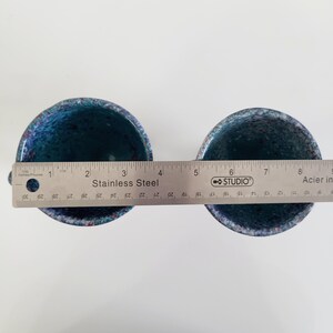 May include: Two speckled blue ceramic cups with a stainless steel ruler placed across them. The ruler measures in inches and centimeters. The cups have a dark blue interior and a speckled exterior with hints of red.