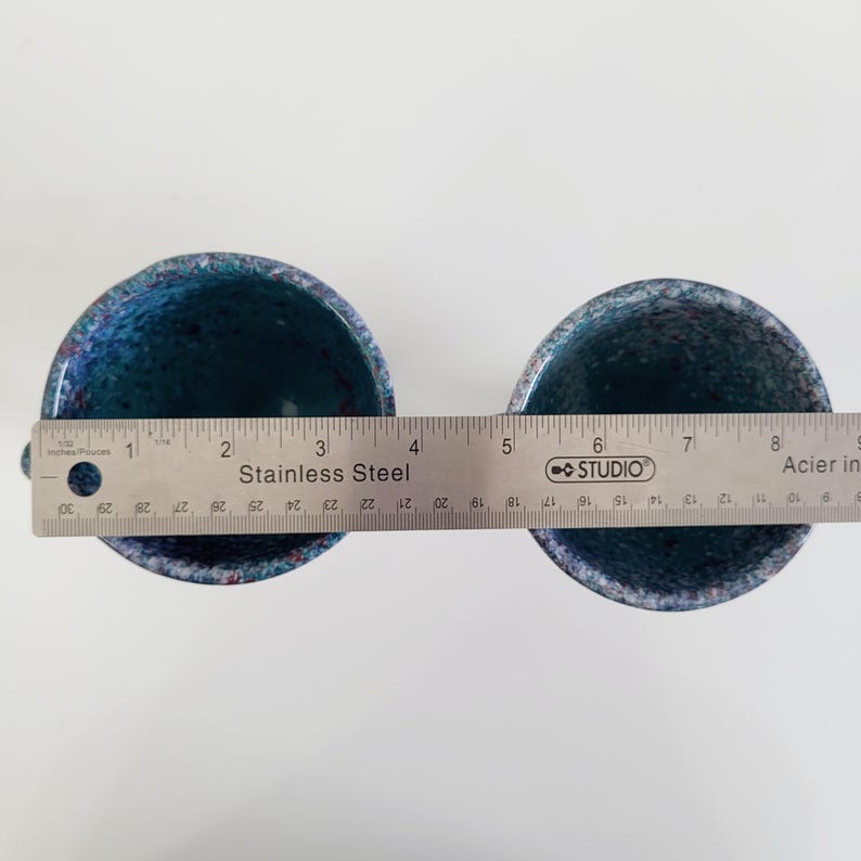 May include: Two speckled blue ceramic cups with a stainless steel ruler placed across them. The ruler measures in inches and centimeters. The cups have a dark blue interior and a speckled exterior with hints of red.