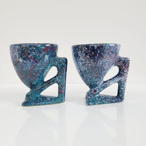 May include: Two speckled ceramic mugs. The mugs are teal, blue, and red with a unique handle design. The handles have a geometric cutout. The mugs have a speckled pattern and a glossy finish. The mugs are on a white surface.