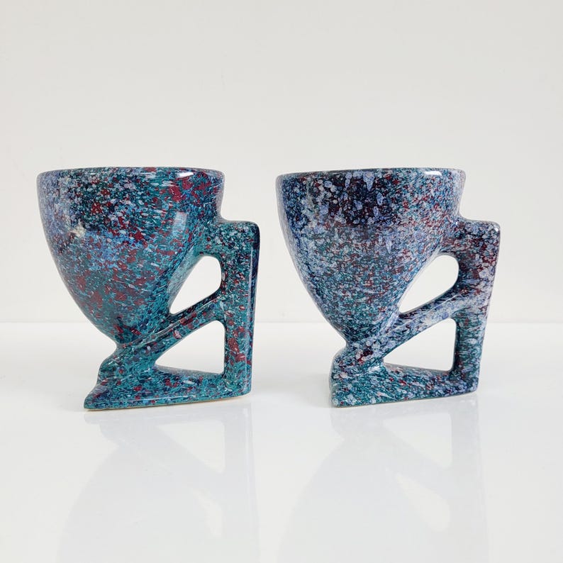 May include: Two speckled ceramic mugs. The mugs are teal, blue, and red with a unique handle design. The handles have a geometric cutout. The mugs have a speckled pattern and a glossy finish. The mugs are on a white surface.