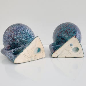 May include: Two ceramic bookends. Each bookend features a speckled blue and teal sphere atop a triangular base. The sphere has red accents. The base is off-white with a hole and etched lines. The bookends are on a white surface.