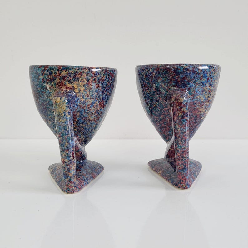 May include: Two speckled ceramic cups with handles. The cups have a unique shape, with a wide top that tapers down to a narrow base. The speckled pattern includes blue, red, and yellow colors. The handles are curved and attached to the side of the cup.